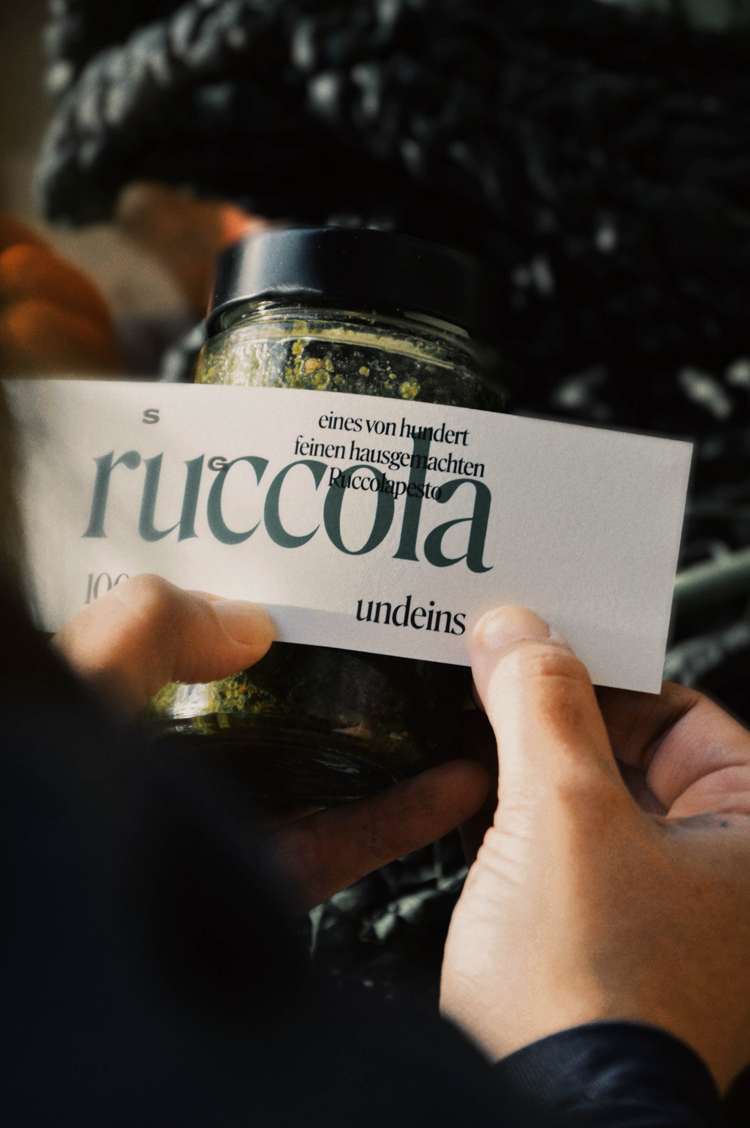 Das Bild zeigt, wie Simone gerade ein Etikett auf das Ruccola Pestoglas klebt. Auf dem Etikett steht in großen, dunkelgrünen Buchstaben Ruccola. Darüber in schwarzer Schrift eines von und feinen hausgemachten Ruccolapesto. Darunter der Markennamen 100undeins. Man sieht Simones Hände.